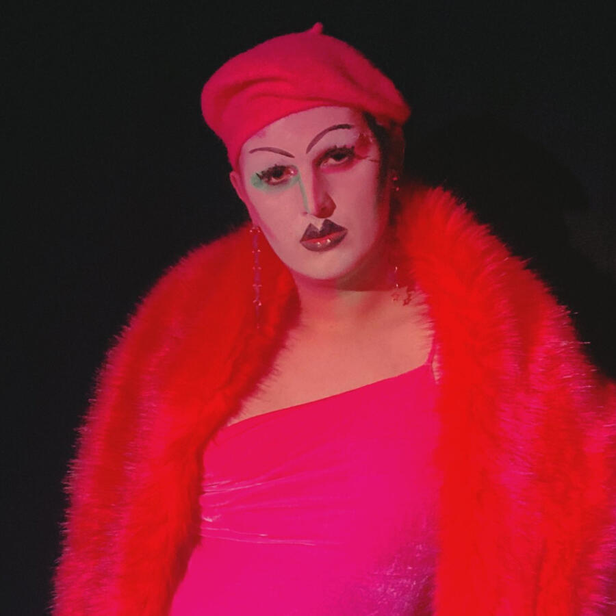 The drag artist, Pop Tart, with striking white make-up, pink dress, red faux-fur shawl, and red beret, posing for a lip-sunc smackdown event.