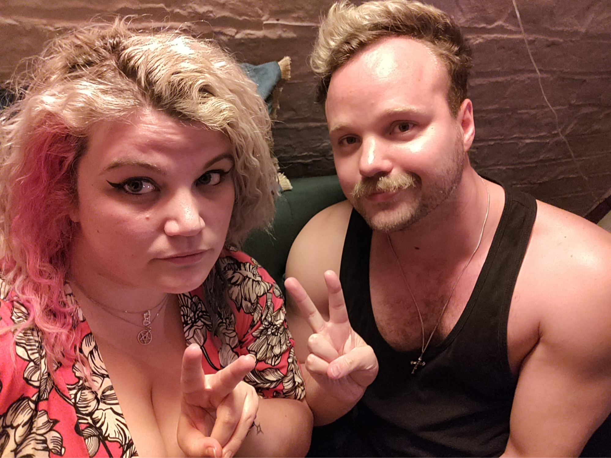 Selfie of Rosie Fox and James Greenwood-Reeves in the corner of a bar, smiling and giving peace signs. Rosie has platinum and pink wavy hair, winged eyeliner, and a coloureful floral dress. James has dyed blonde hair, a big gay moustasche, and is wearing a